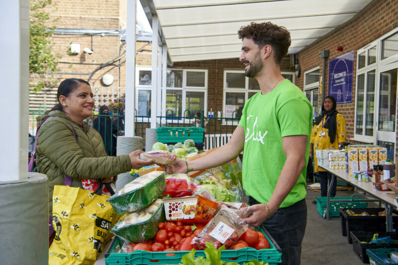 Man in green t-shirt handing food to woman in a winter jacket at a table of fruit and vegetables